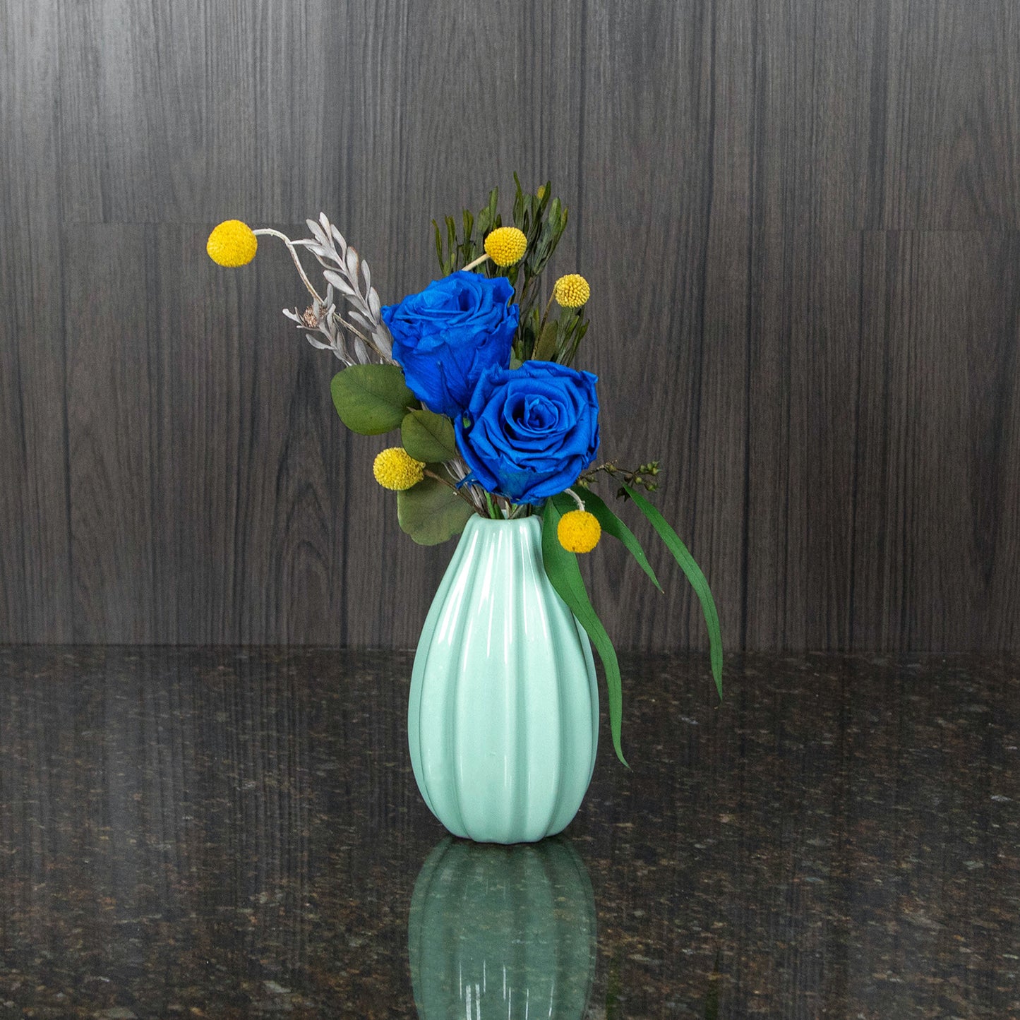 a mint colored ceramic bud vase with two blue preserved roses, dried greenery, and dried craspedia