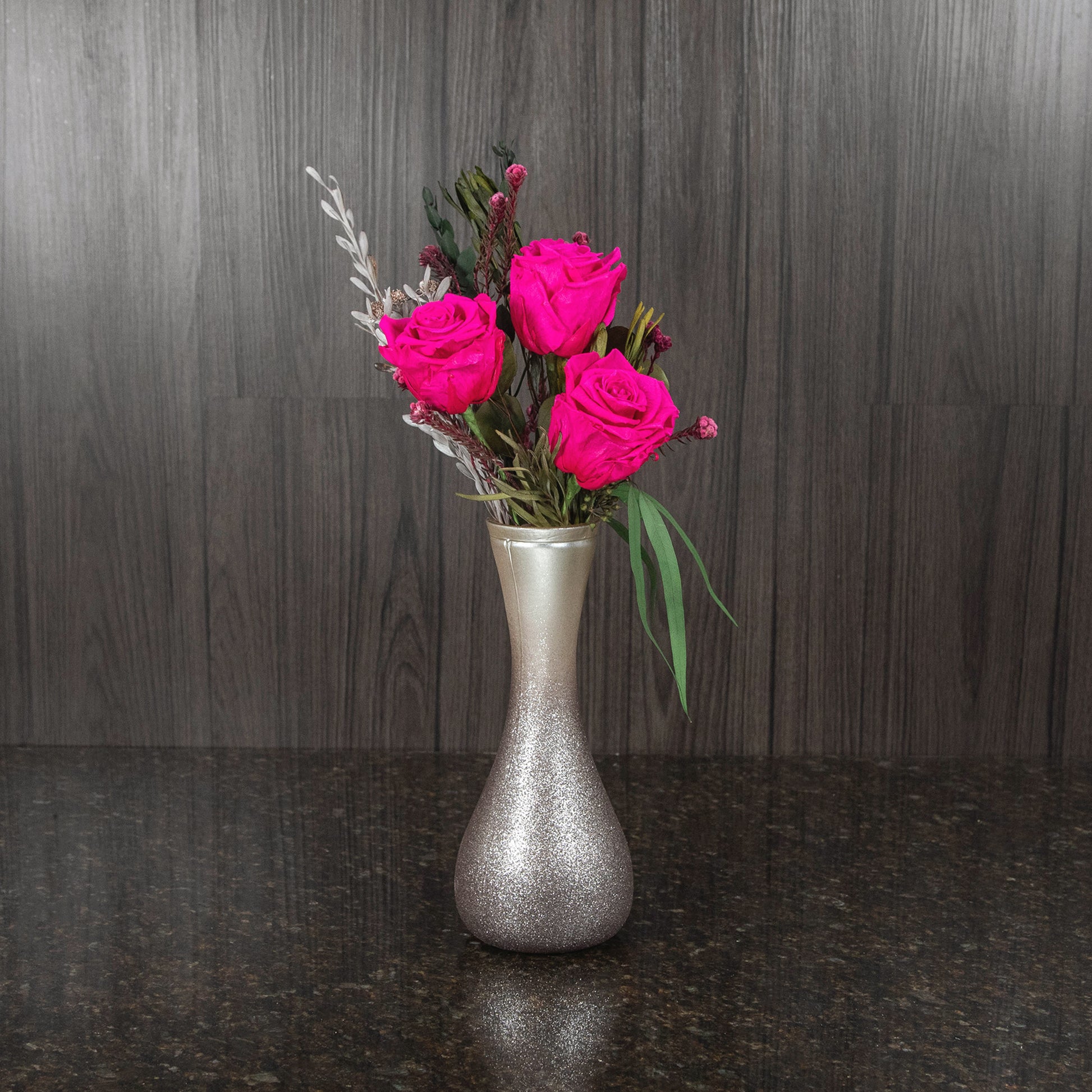 three magenta preserved roses and dried greenery in a tall sparkly champagne colored vase