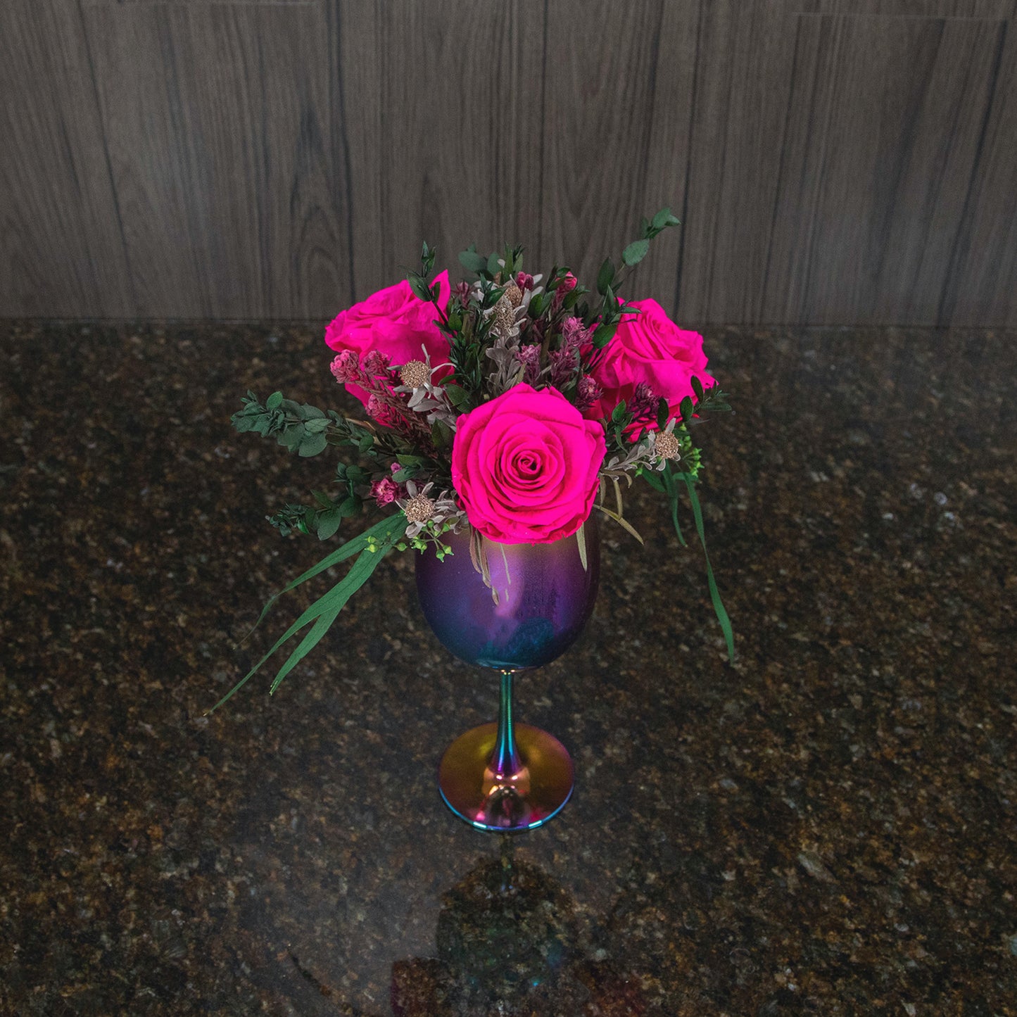 top down view of three magenta preserved roses and dried greenery in a colorful metallic vase