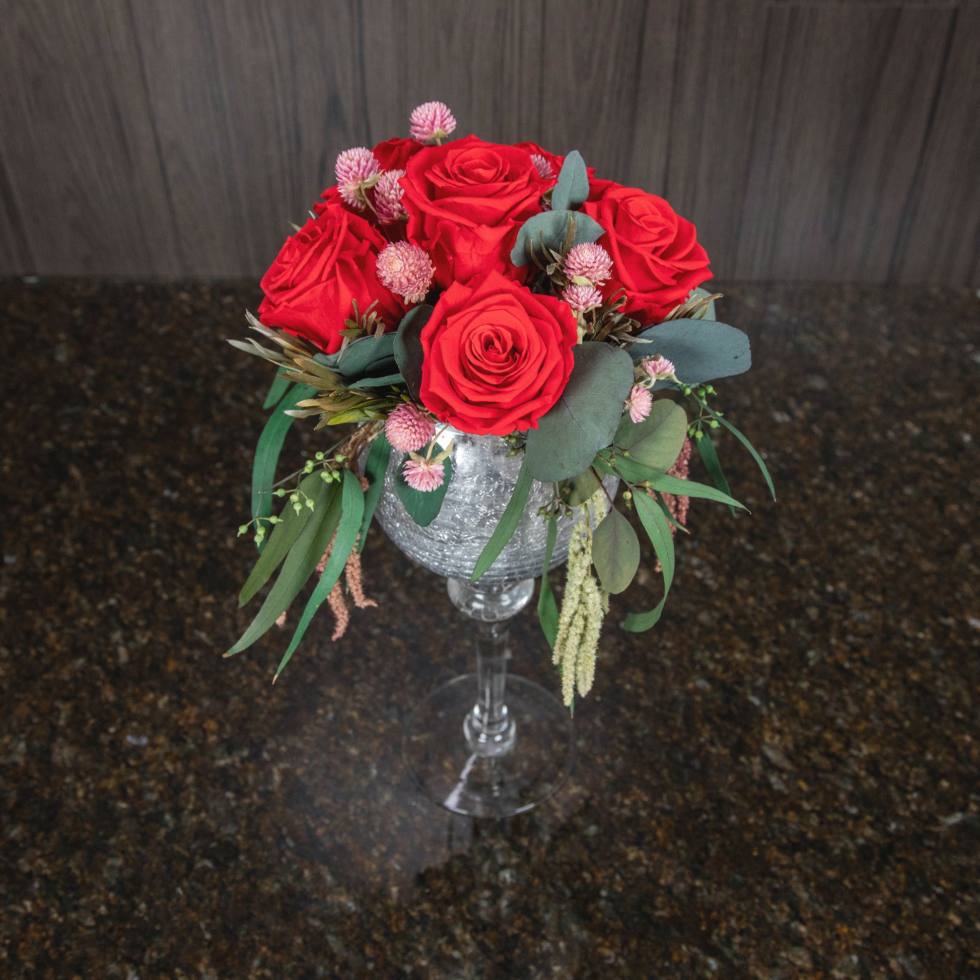 top down view of 6 preserved red roses and dried greenery in a tall silver stemmed glass vase