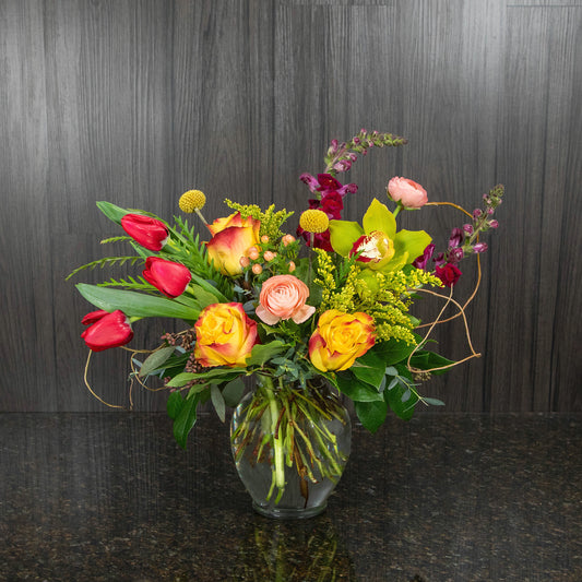 a whimsical mix of warm colored flowers in an urn shaped glass vase