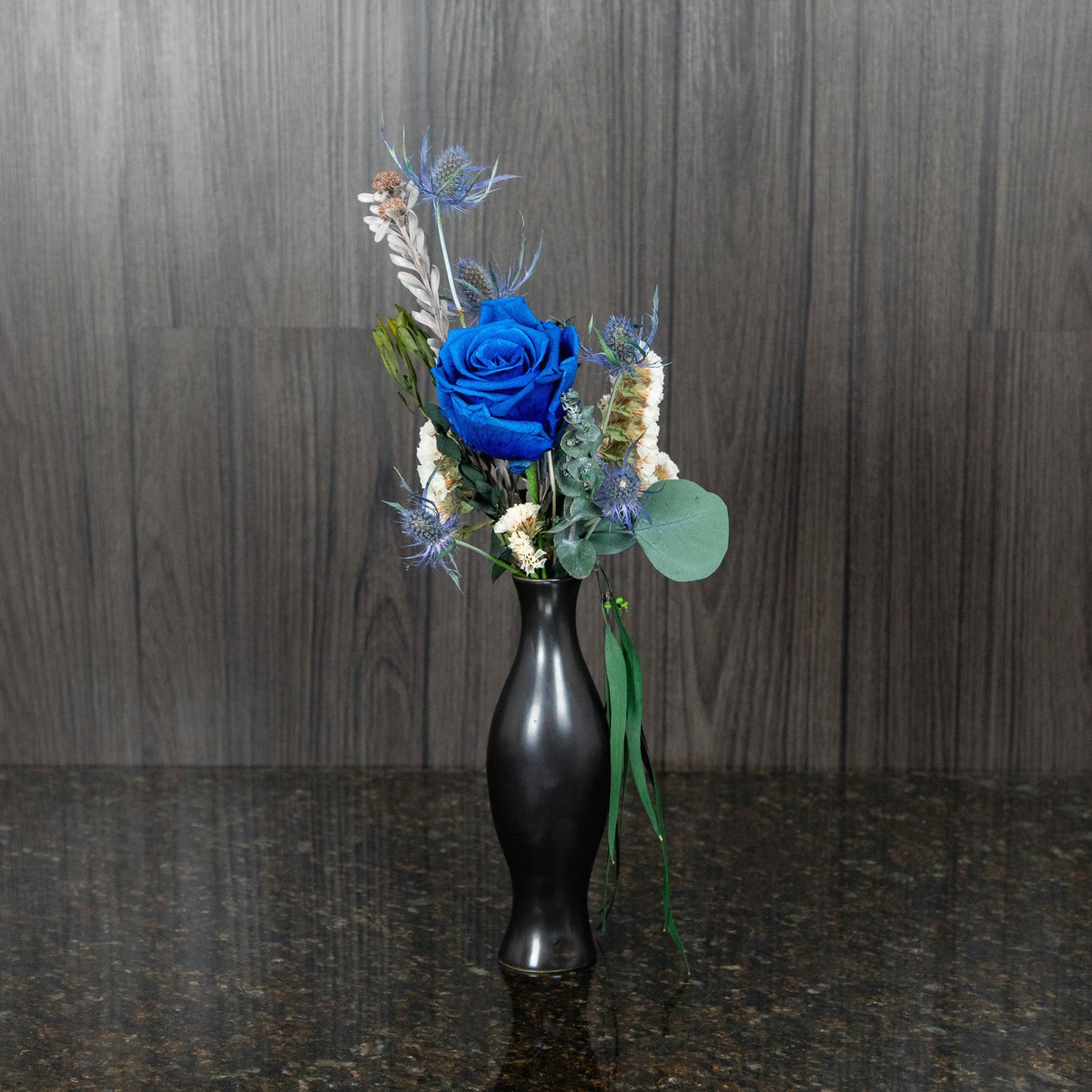 a single blue preserved rose, dried greenery, and dried thistle in a black ceramic bud vase