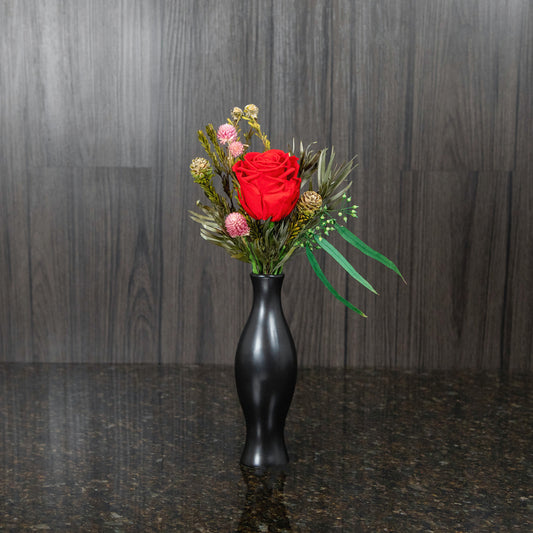 a single red preserved rose and greenery in a black bud vase