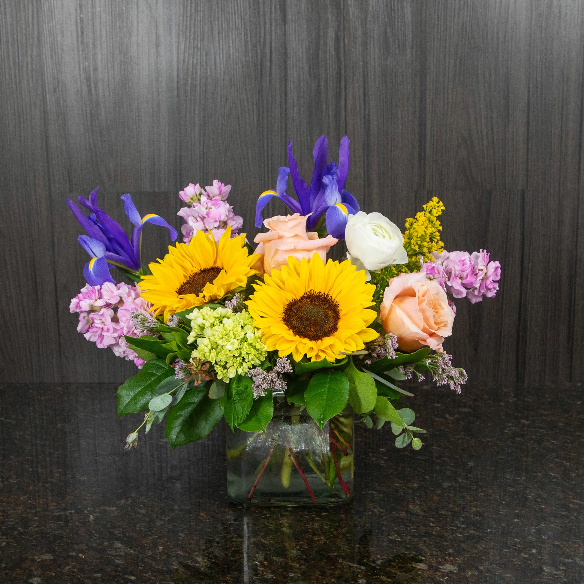 a colorful flower arrangement with sunflowers and other cheerful flowers in a glass cube vase