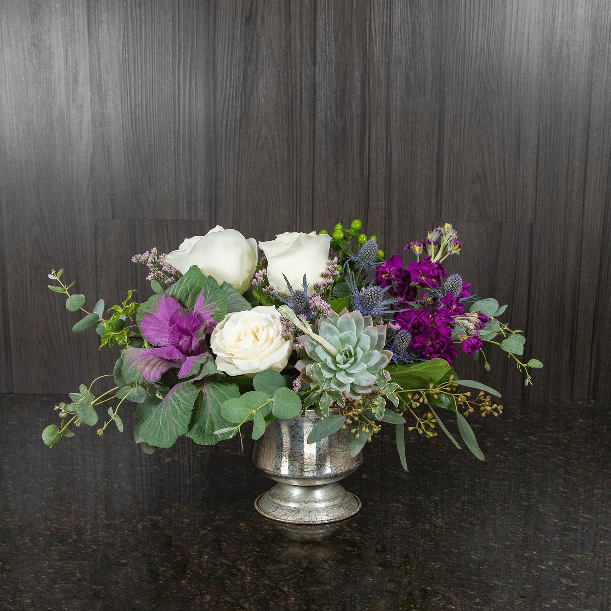 a flower arrangement in whites, purples, greens, and blues in a silver metal urn vase